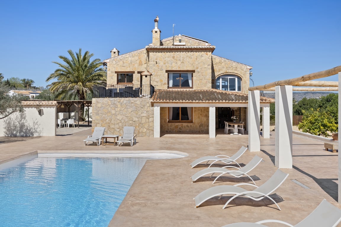 Natursteinfassade mit überdachter Naya, weitläufiger Fliesenterrasse und Pool. Die Villa präsentiert ihre zweistöckige Architektur und sonnige Außenliegebereiche.