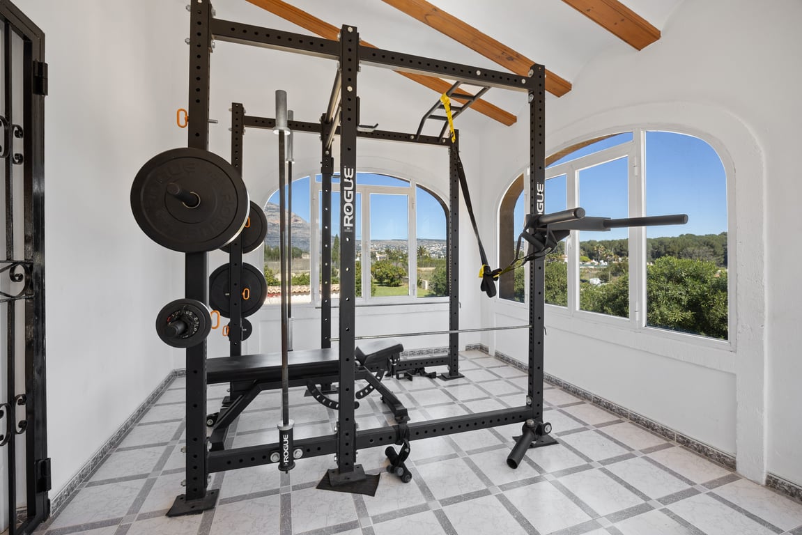 Privater Fitnessraum mit professionellem Rogue-Equipment, untergebracht in einem hellen Raum mit Holzbalkendecke und weitem Blick auf die Landschaft von Jávea.