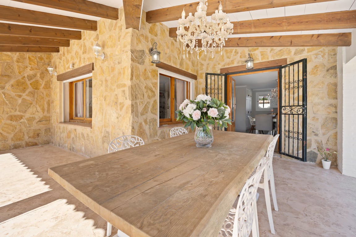 Überdachte Terrasse mit Natursteinwänden, Sichtbalken aus Holz und einem großen Esstisch unter einem klassischen Kronleuchter.