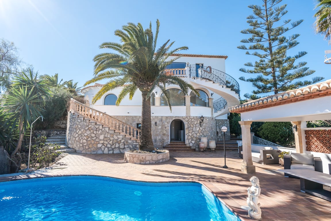 Außenansicht der Villa mit Pool, Natursteinfassade, geschwungenen Treppenaufgängen und einem mediterranen Garten mit Palmen unter klarem Himmel.