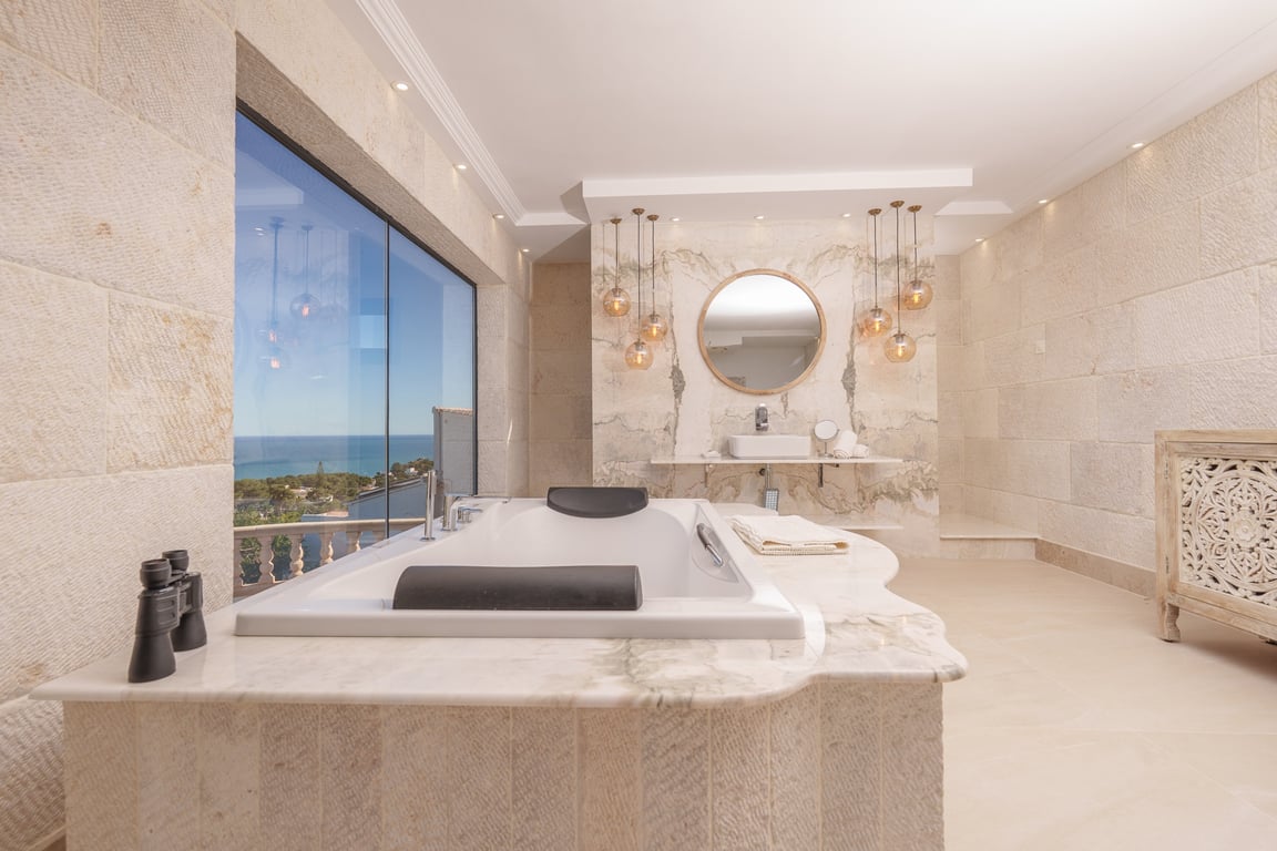 Badezimmer der Master-Suite mit Kalksteinwänden, in Marmor eingefasster Badewanne mit Meerblick und Designer-Waschtisch mit dekorativen Pendelleuchten.