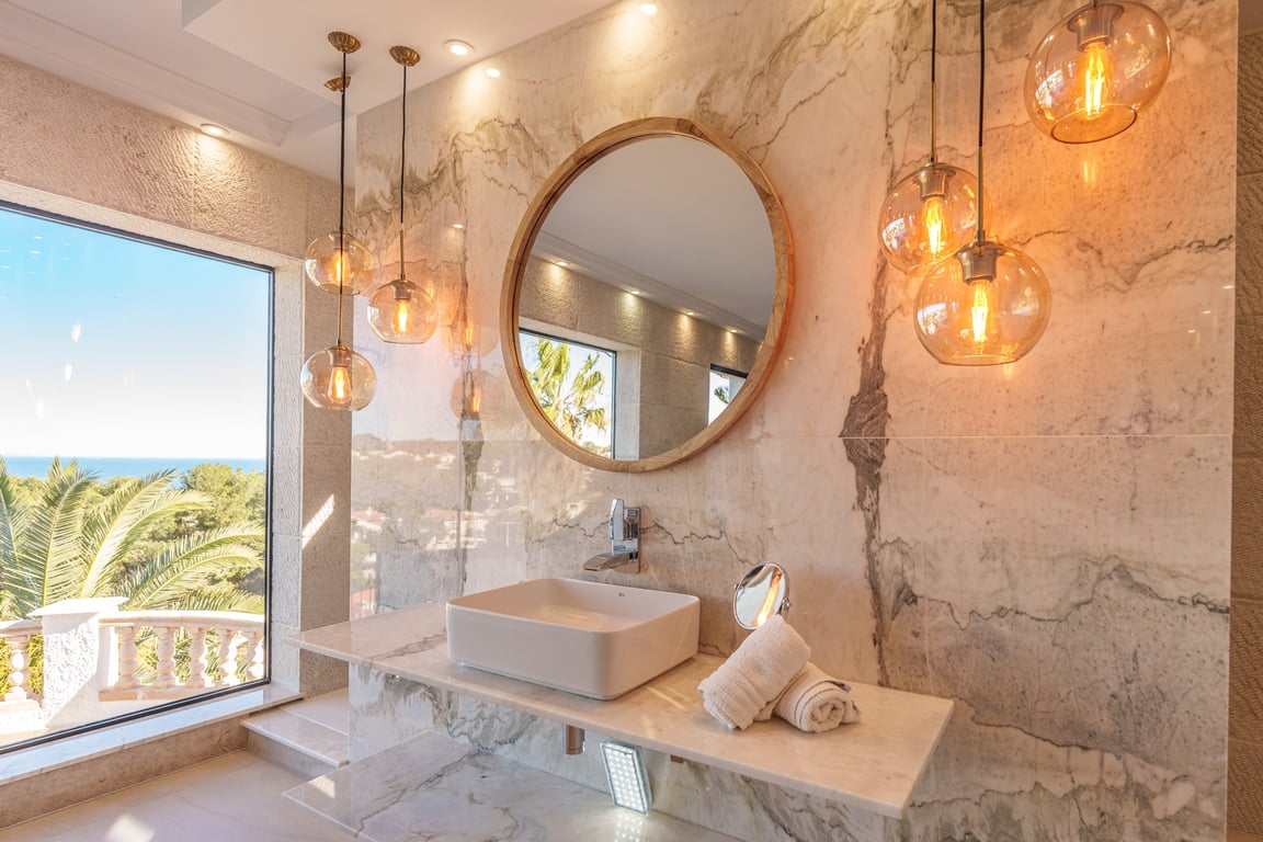 Badezimmer der Master-Suite mit marmorierten Wänden, Aufsatzwaschbecken, rundem Holzspiegel und Glaspendelleuchten mit Blick auf das Mittelmeer.