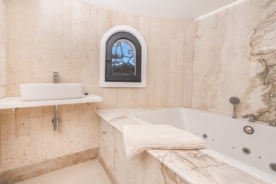 Badezimmer mit marmorverkleideter Whirlpool-Badewanne, Aufsatzwaschbecken und Wänden aus Naturstein mit einem charakteristischen Rundbogenfenster.