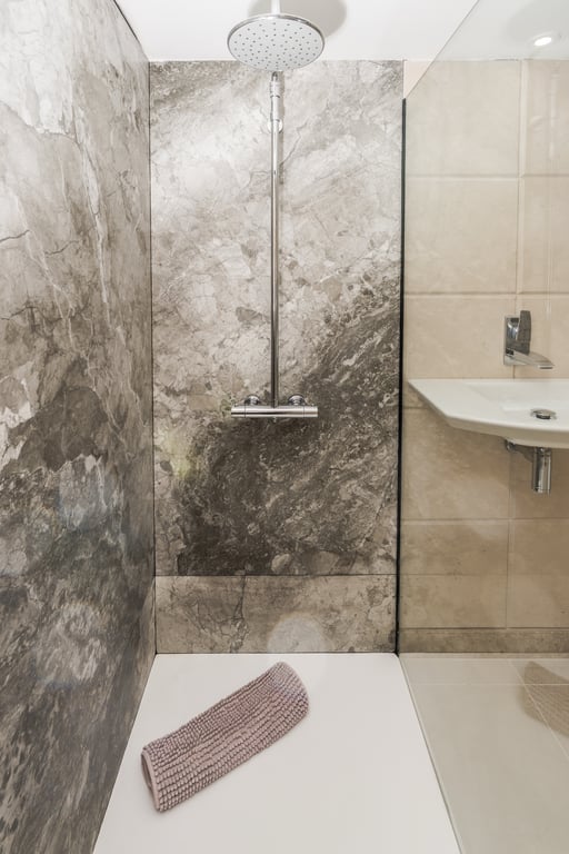 Begehbare Dusche mit grauen Marmorfliesen, verchromtem Regenduschkopf und weißer Duschwanne, abgetrennt durch eine Glaswand.