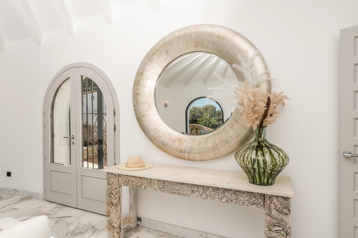 Eingangsbereich mit Marmorboden, geschnitzter Holzkonsole und großem Rundspiegel. Weiße Balkendecken und Rundbogentüren unterstreichen das elegante mediterrane Design.