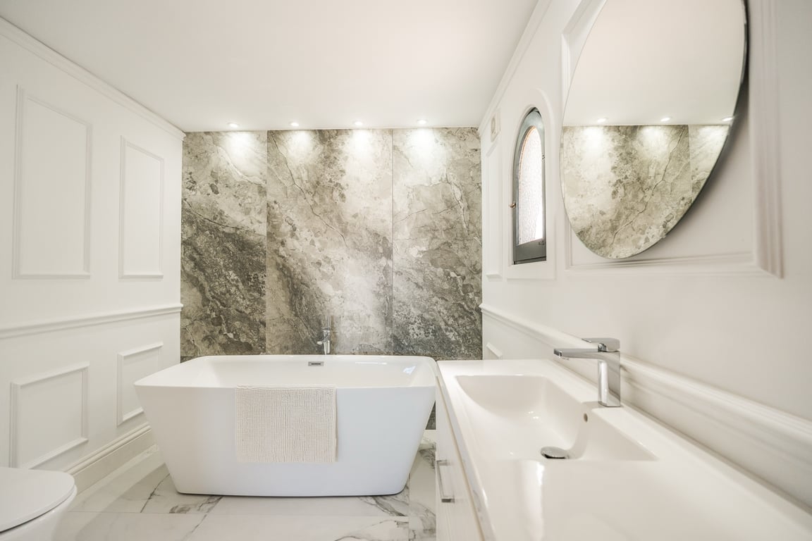 Elegantes Badezimmer mit freistehender Badewanne, Marmor-Akzentwand mit Spotlights und klassischen weißen Wandpaneelen.