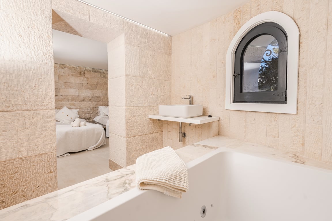 En-Suite-Badezimmer mit Natursteinwänden, einer Badewanne mit Marmoreinfassung und modernem Waschbecken, das direkt an das Schlafzimmer mit Rundbogenfenster angrenzt.