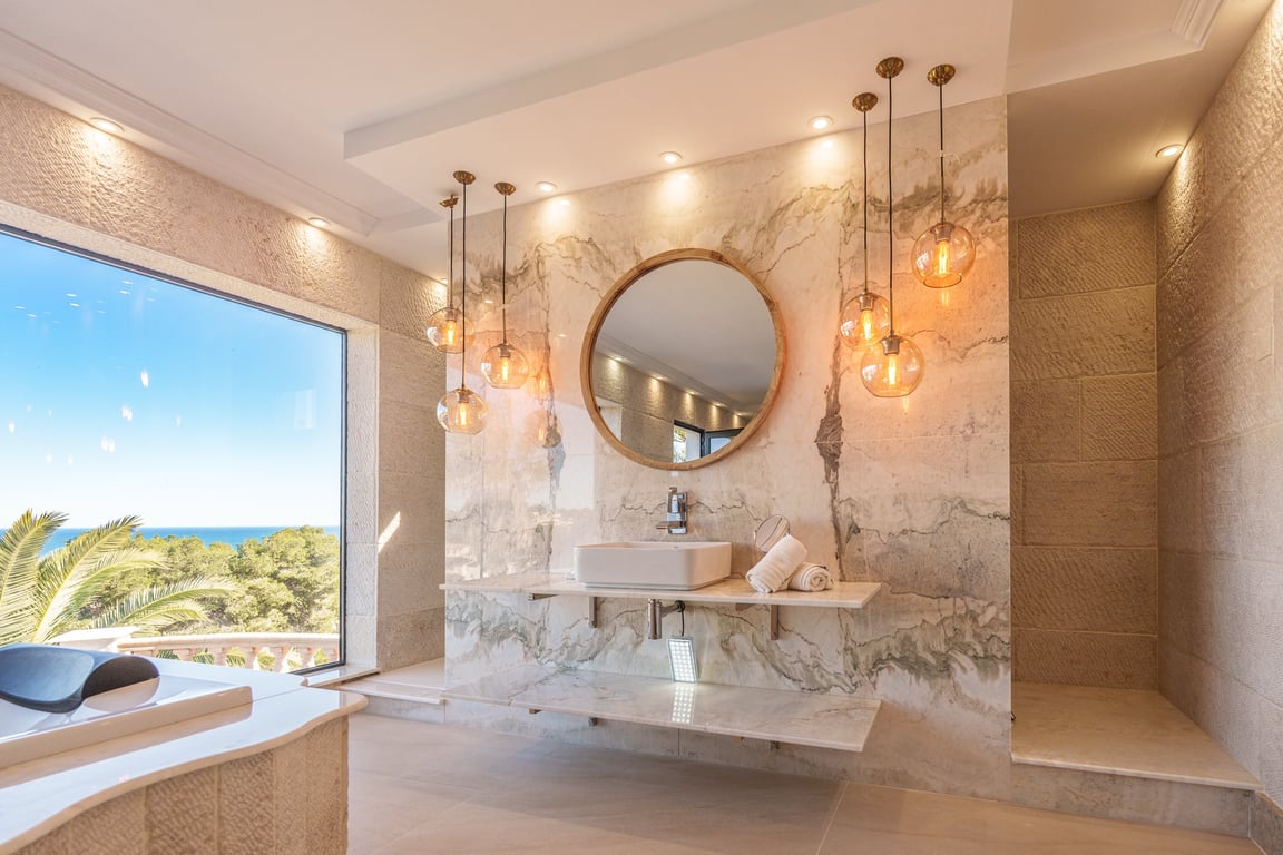 Hauptbadezimmer mit gemaserten Marmorwänden, Aufsatzwaschbecken auf schwebendem Waschtisch und Rundspiegel. Glaskugelleuchten und ein großes Fenster bieten einen weiten Meerblick.