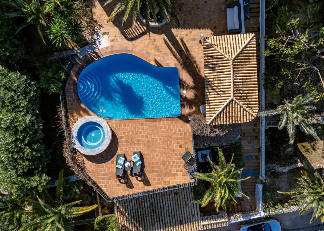 Luftaufnahme des Außenbereichs mit nierenförmigem Pool, rundem Whirlpool und weitläufiger Terrasse, umgeben von Palmen und mediterraner Bepflanzung.