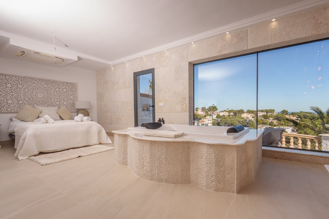 Master-Suite mit integrierter Naturstein-Badewanne, großen Panoramafenstern mit Meerblick und einer hellen, offenen Raumgestaltung.
