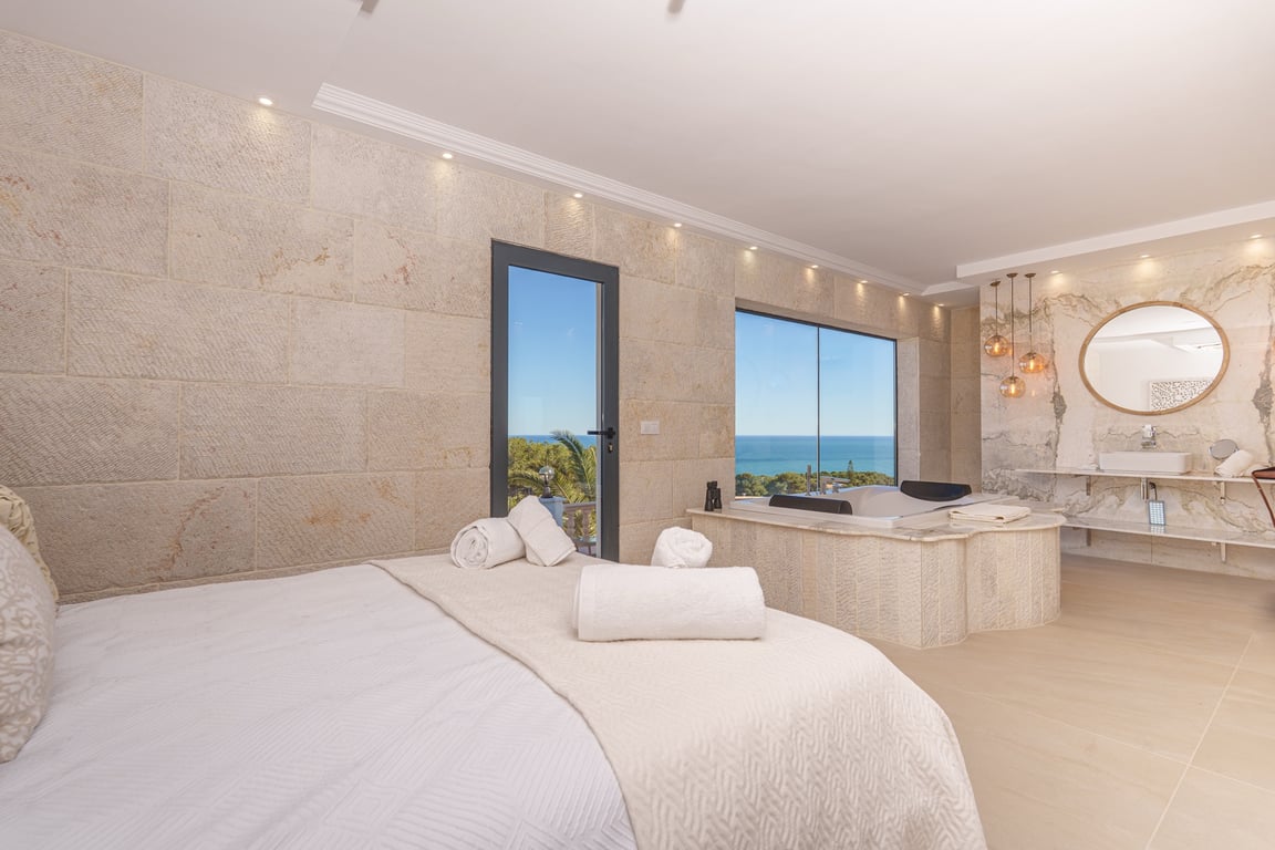 Master-Suite mit Natursteinwänden, integriertem Whirlpool am Panoramafenster mit Meerblick und offenem Badezimmer mit Marmorelementen.