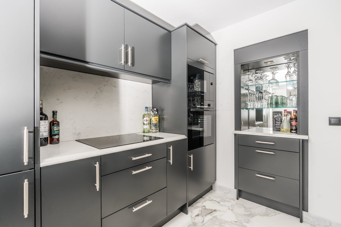 Moderne Küche mit anthrazitfarbenen Schränken, Arbeitsplatten aus weißem Marmor und integrierten Geräten. Ein Bar-Bereich mit beleuchteter Glasvitrine ergänzt den Marmorboden.