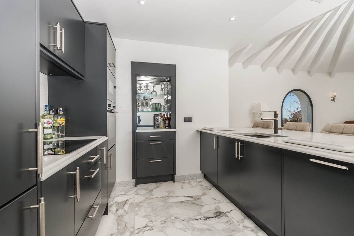 Moderne Küche mit dunklen Fronten, Marmoroberflächen und einer zentralen Insel. Ein integriertes Barfach und die markante Balkendecke ergänzen das elegante Design.
