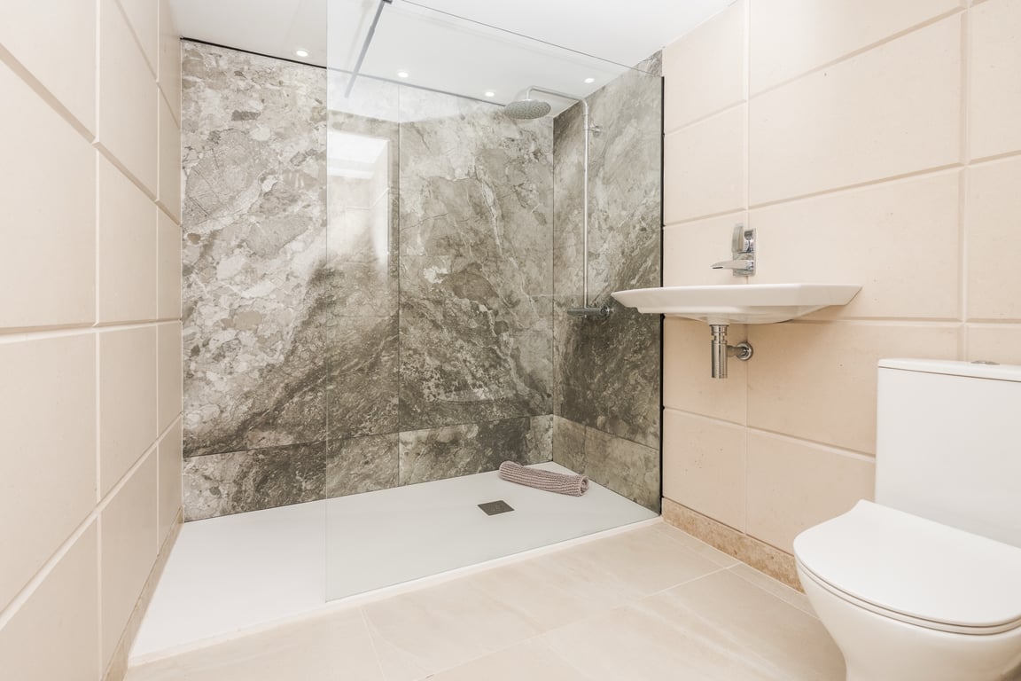 Modernes Badezimmer mit begehbarer Dusche, Glastrennwand und Wand in Marmoroptik. Ausgestattet mit weißen Sanitärobjekten und cremefarbenen Fliesen.