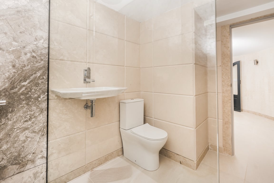 Modernes Badezimmer mit cremefarbenen Kalksteinfliesen, zeitgemäßem Stand-WC und Händewaschbecken. Ausgestattet mit Glasduschwand und Marmorakzenten an der Wand.