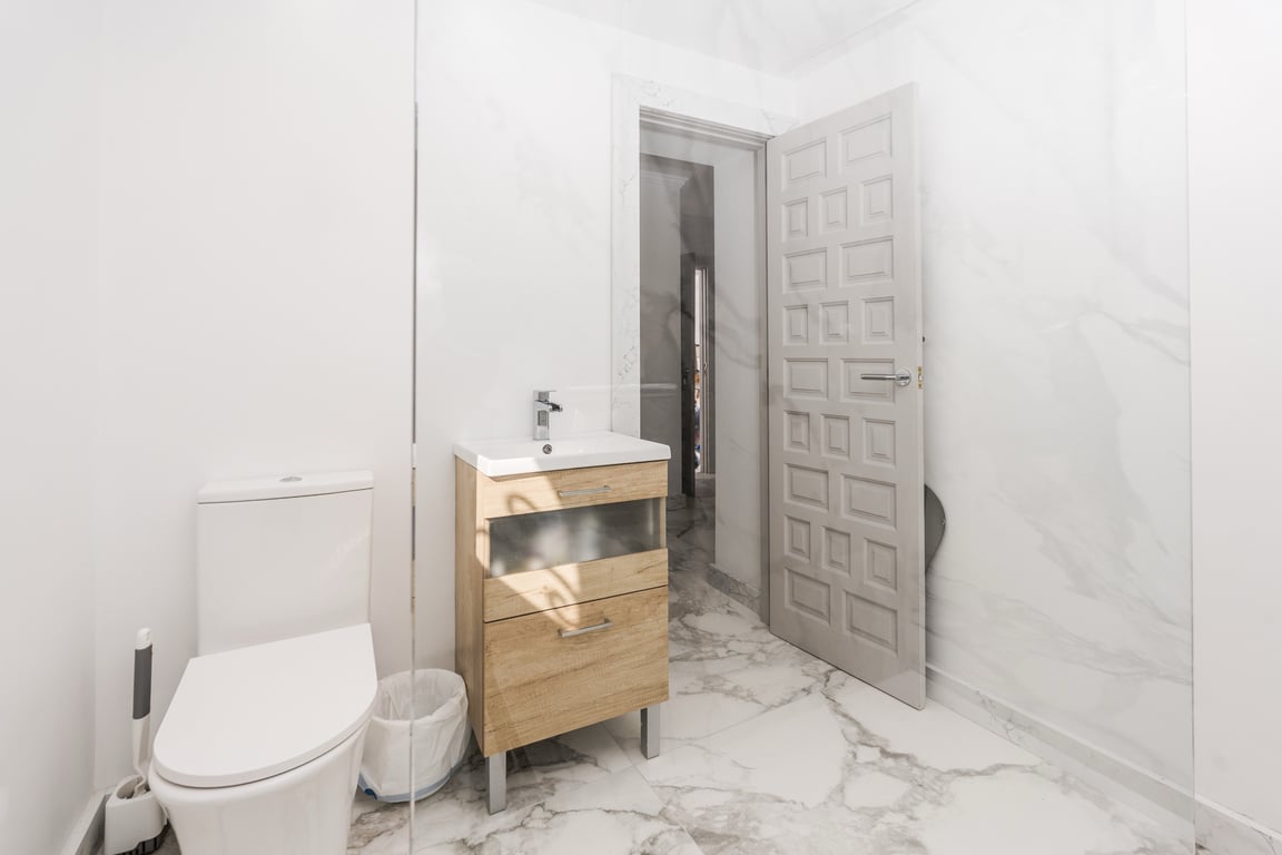 Modernes Badezimmer mit weißen Marmoreffekt-Fliesen, einem Waschbeckenunterschrank aus hellem Holz und einer festen Glasduschwand.