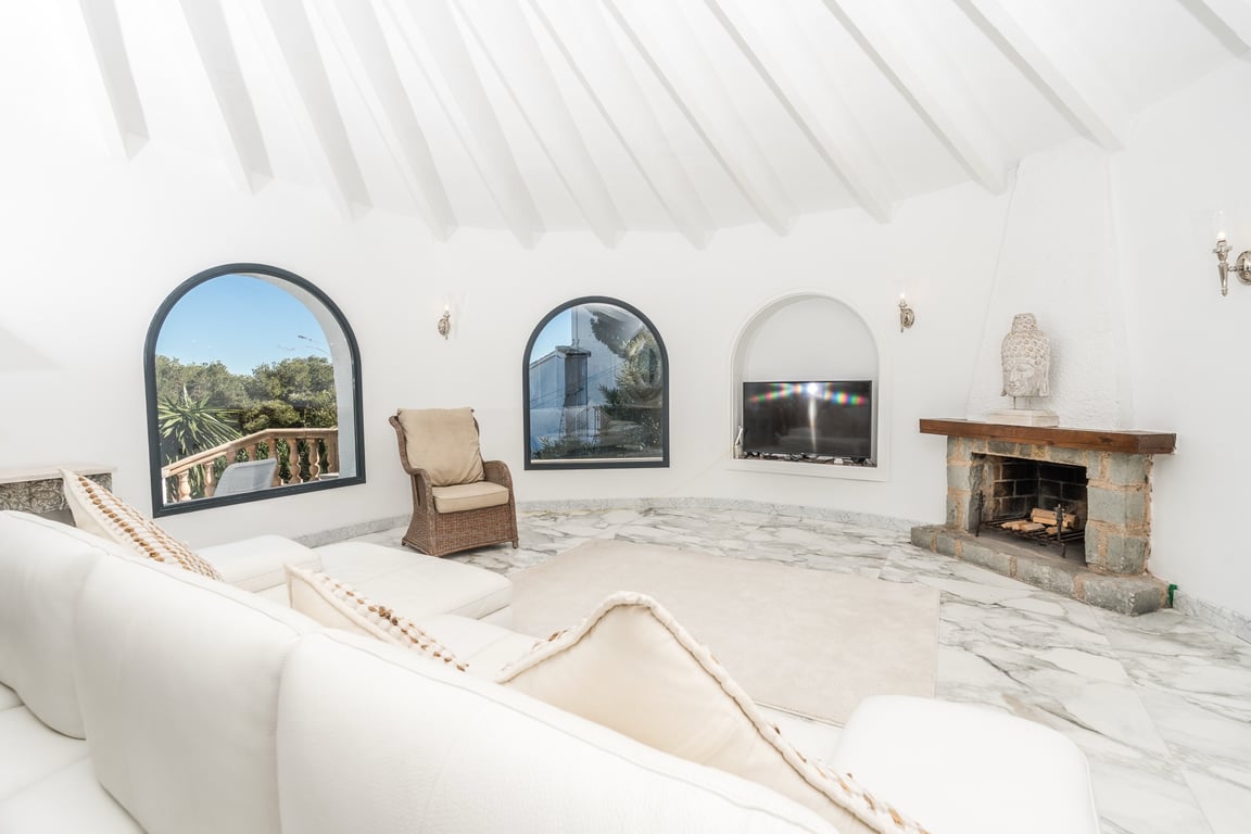 Rundes Wohnzimmer mit weißer Balkendecke, Marmorboden und Natursteinkamin. Rundbogenfenster lassen viel Tageslicht herein und bieten Ausblicke ins Grüne.