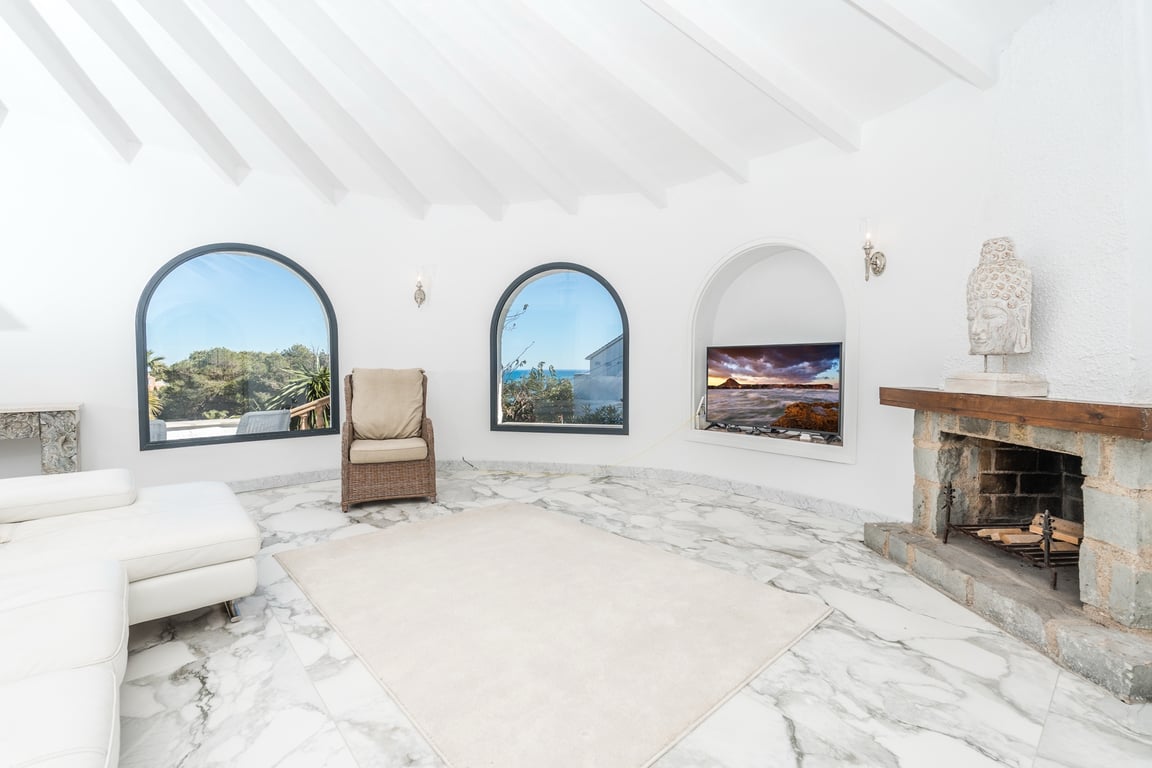 Rundes Wohnzimmer mit weißer Balkendecke, Marmorboden und rustikalem Steinkamin. Rundbogenfenster bieten Ausblicke auf das Meer und die umliegende Natur.