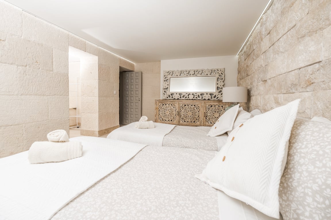 Schlafzimmer mit Wänden aus Kalksteinblöcken, zwei Einzelbetten und geschnitzten Holzmöbeln. Indirekte LED-Deckenbeleuchtung betont die Steintextur.