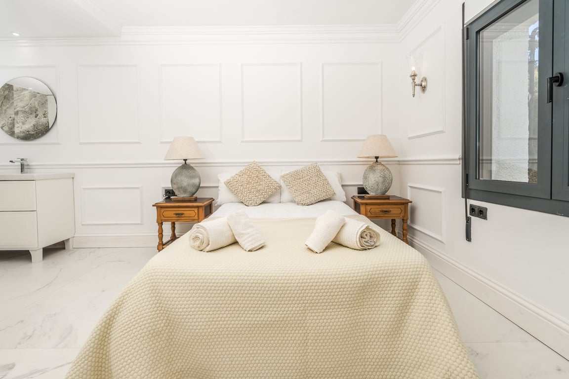 Schlafzimmer mit weißen Wandpaneelen und Marmorboden. Es verfügt über einen integrierten Waschtisch mit Rundspiegel und klassische Nachttische aus Holz.