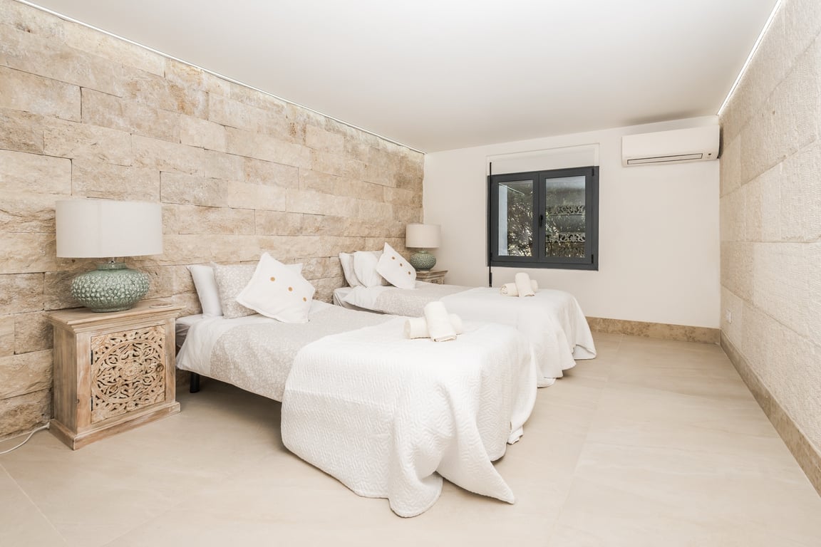 Schlafzimmer mit zwei Einzelbetten, einer Wand aus strukturiertem Naturstein, geschnitzten Nachttischen aus Holz und integrierter LED-Deckenbeleuchtung.