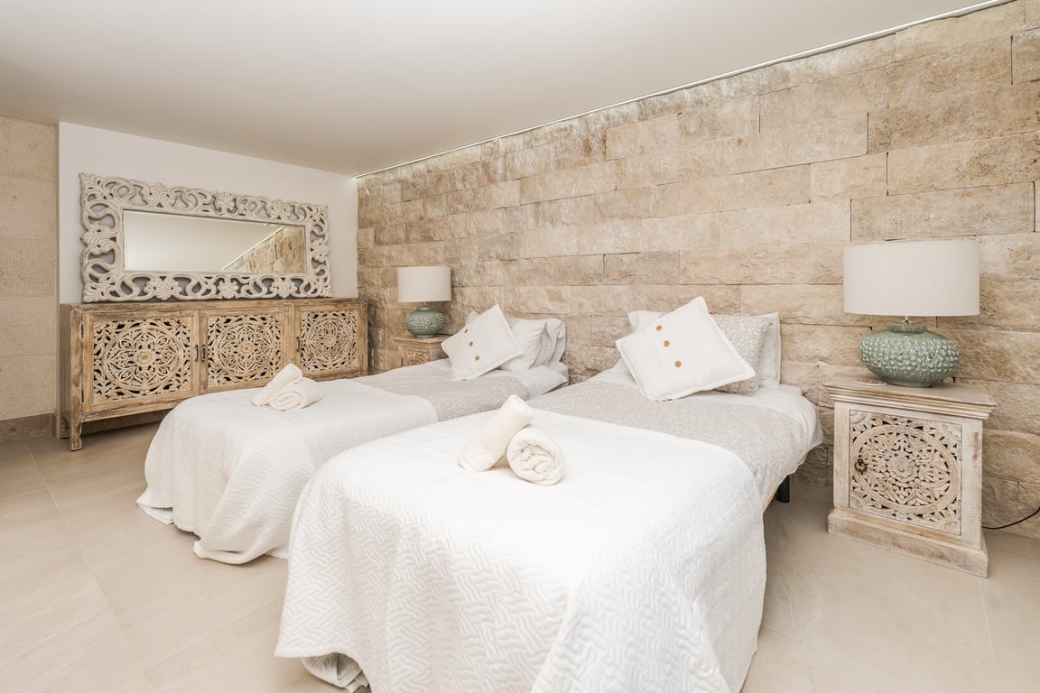 Schlafzimmer mit zwei Einzelbetten, einer Wand aus strukturiertem Naturstein und handgeschnitzten Holzmöbeln. Integrierte Deckenbeleuchtung und helle Steinböden.