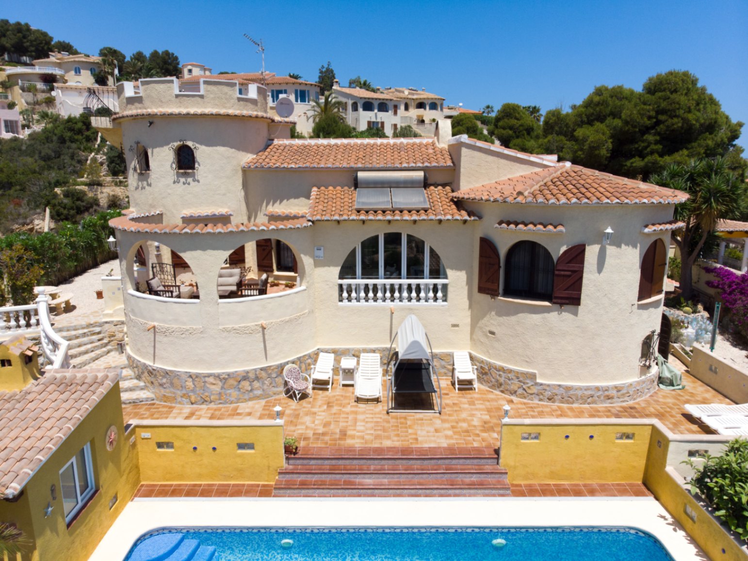 Außenansicht der Villa mit markantem Turm, Bogenterrassen und Pool. Zu sehen sind mediterrane Ziegeldächer mit Solaranlage, Holzschlagläden und eine Natursteinfassade im Sockelbereich.
