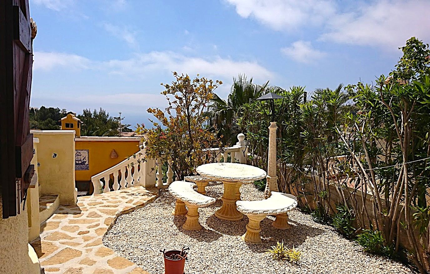 Außenbereich mit einem runden Steintisch und verzierten Bänken auf Kies und Natursteinpflaster, umgeben von mediterranen Pflanzen und Meerblick.