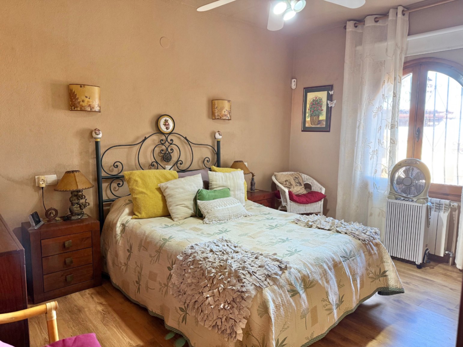 Rustikales Schlafzimmer mit schmiedeeisernem Kopfteil, Holzboden und Rundbogenfenster. Mit Deckenventilator, Heizkörper und klassischen Nachtschränken.