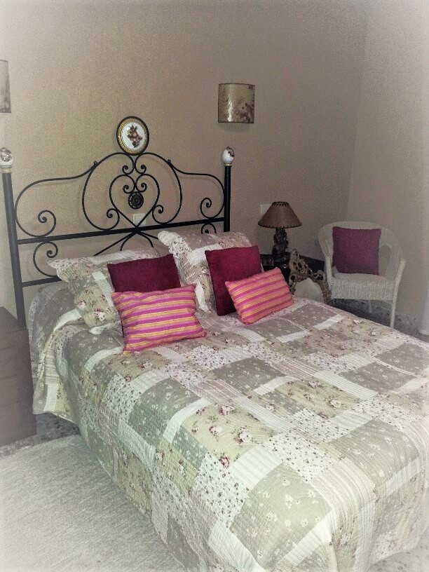 Schlafzimmer mit Doppelbett, schwarzem schmiedeeisernem Kopfteil und Patchwork-Bettwäsche. Ein weißer Korbsessel und neutral gehaltene Wände ergänzen den Raum.
