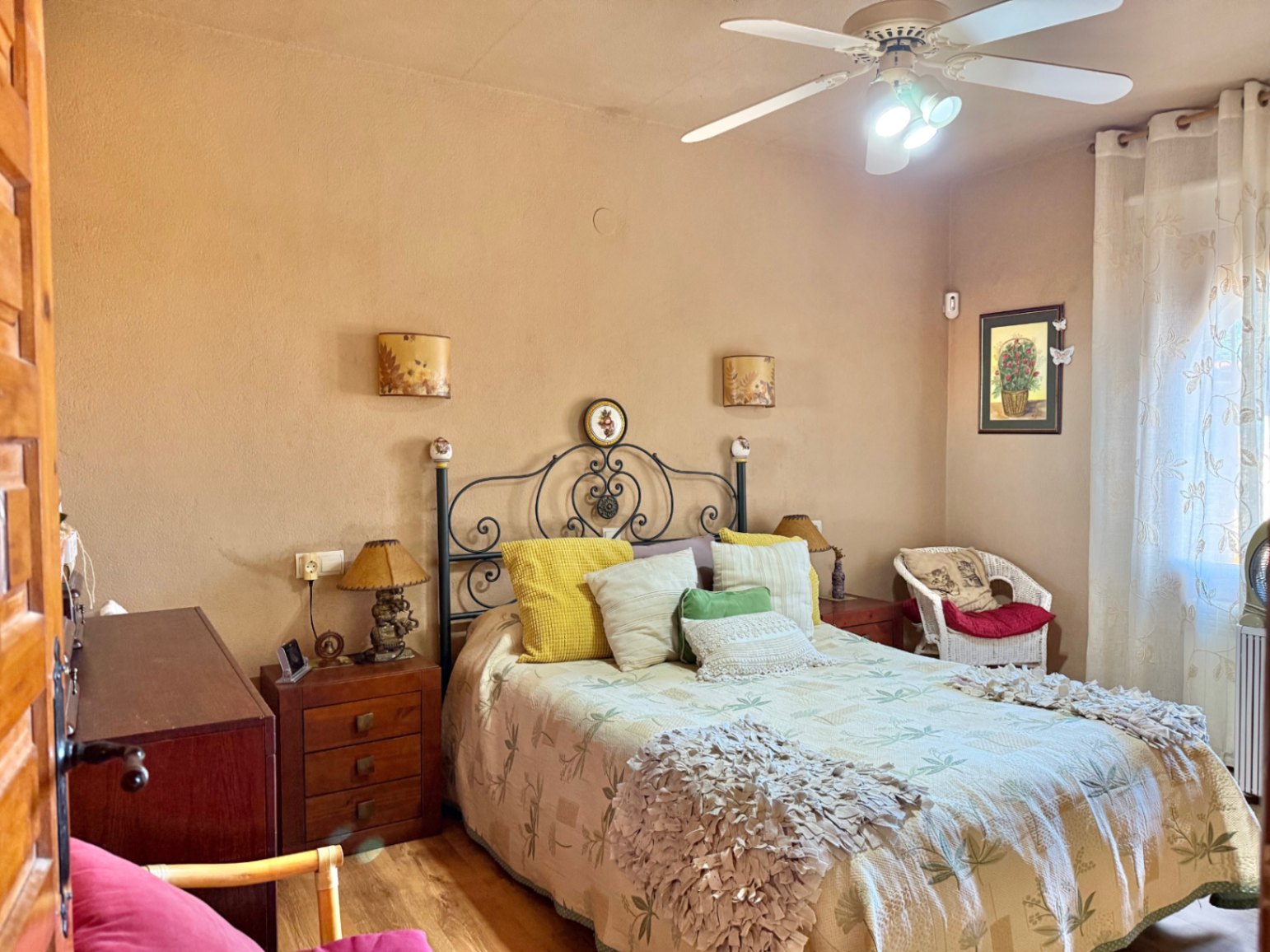 Schlafzimmer mit schmiedeeisernem Kopfteil, Nachttischen aus Holz und Deckenventilator. Ausgestattet mit einem Korbsessel und natürlichem Lichteinfall durch das Fenster.