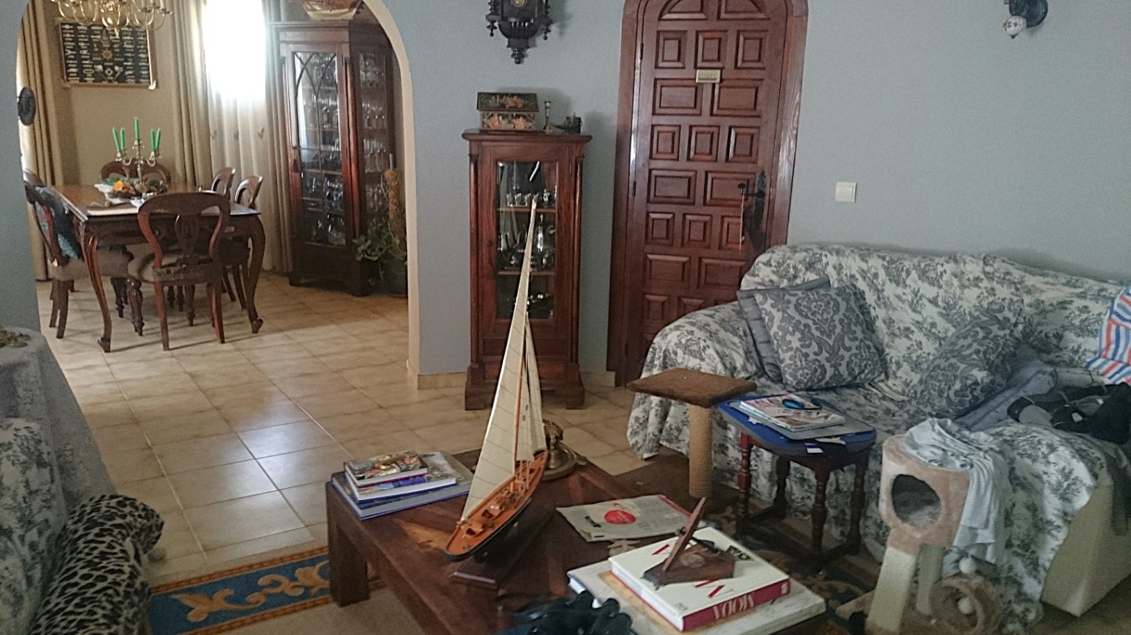 Traditionelles Wohnzimmer mit Keramikfliesen, Rundbogen zum Essbereich und klassischen Holzmöbeln, darunter Vitrinen und ein Couchtisch mit nautischen Dekoelementen.