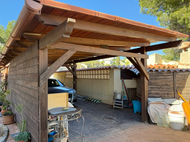 Überdachter Carport aus Holz mit sichtbaren Balken und Ziegeldach, der einen geschützten Stellplatz auf geprägtem Betonboden bietet.