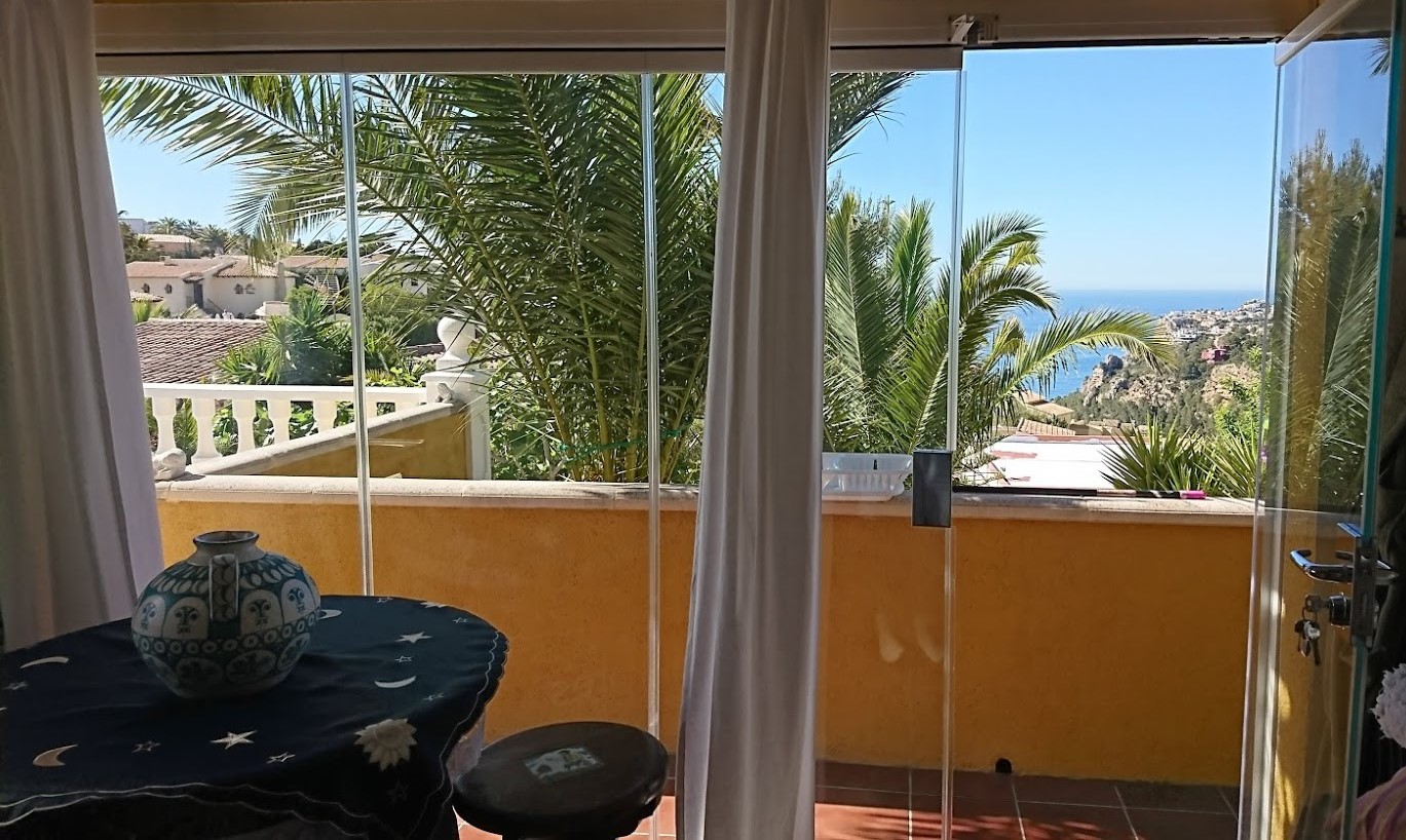 Verglaste Terrasse mit rahmenlosen Glasvorhängen, die einen Blick auf das Mittelmeer und die Palmen im Garten bietet.