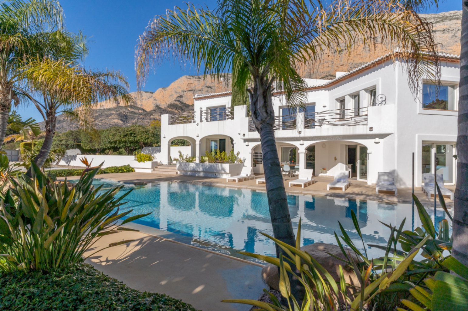 Außenansicht der Villa im Ibiza-Stil mit großem Pool, Rundbogen-Veranden und Terrassen vor der Kulisse des Montgó-Massivs und mediterraner Palmen.