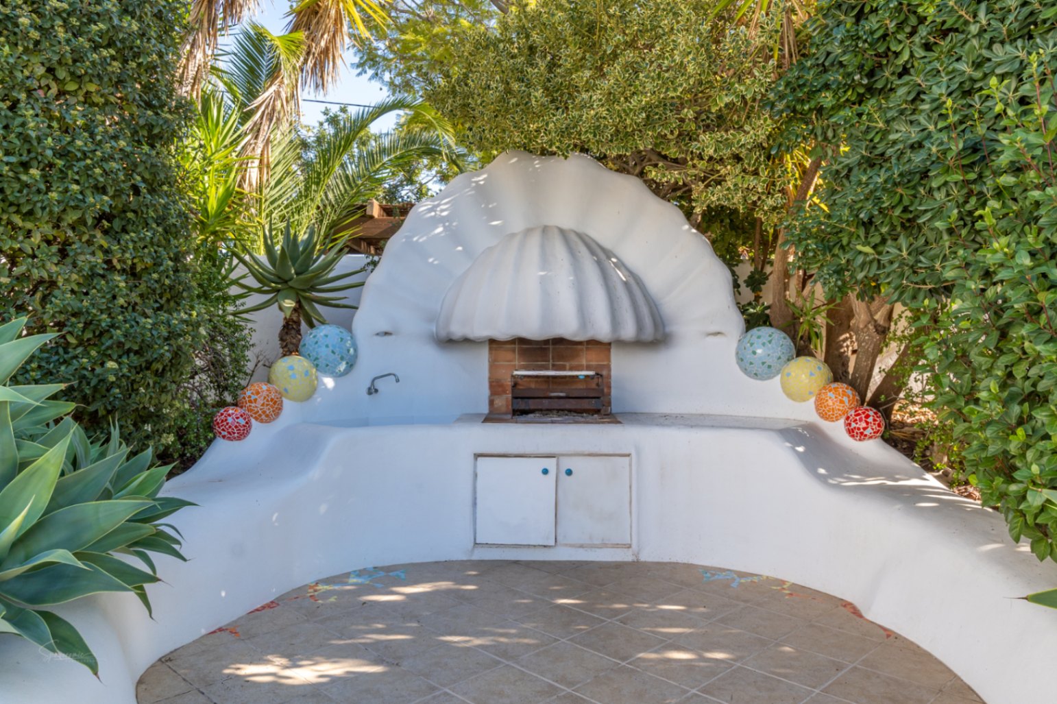 Außengrillbereich im Ibiza-Stil mit einer weißen, muschelförmigen Abzugshaube, geschwungenen gemauerten Arbeitsplatten mit Mosaikdetails und integrierter Spüle im grünen Garten.