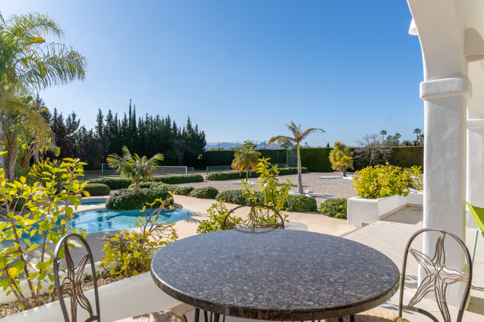 Blick von der Naya mit Granittisch auf den Pool, die mediterrane Gartenanlage und den privaten Tennisplatz unter blauem Himmel.