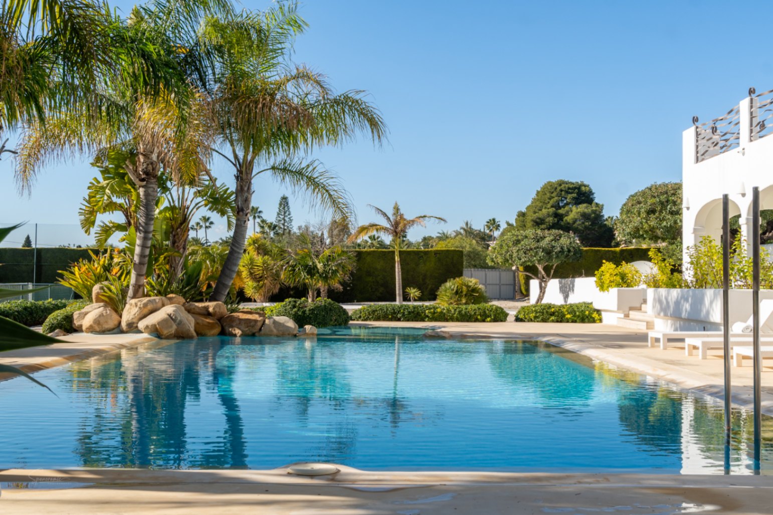 Designer-Pool mit flachem Einstieg, umgeben von tropischen Palmen, Natursteinen und gepflegten Gärten neben der weißen Bogen-Naya und dem Tennisplatz.