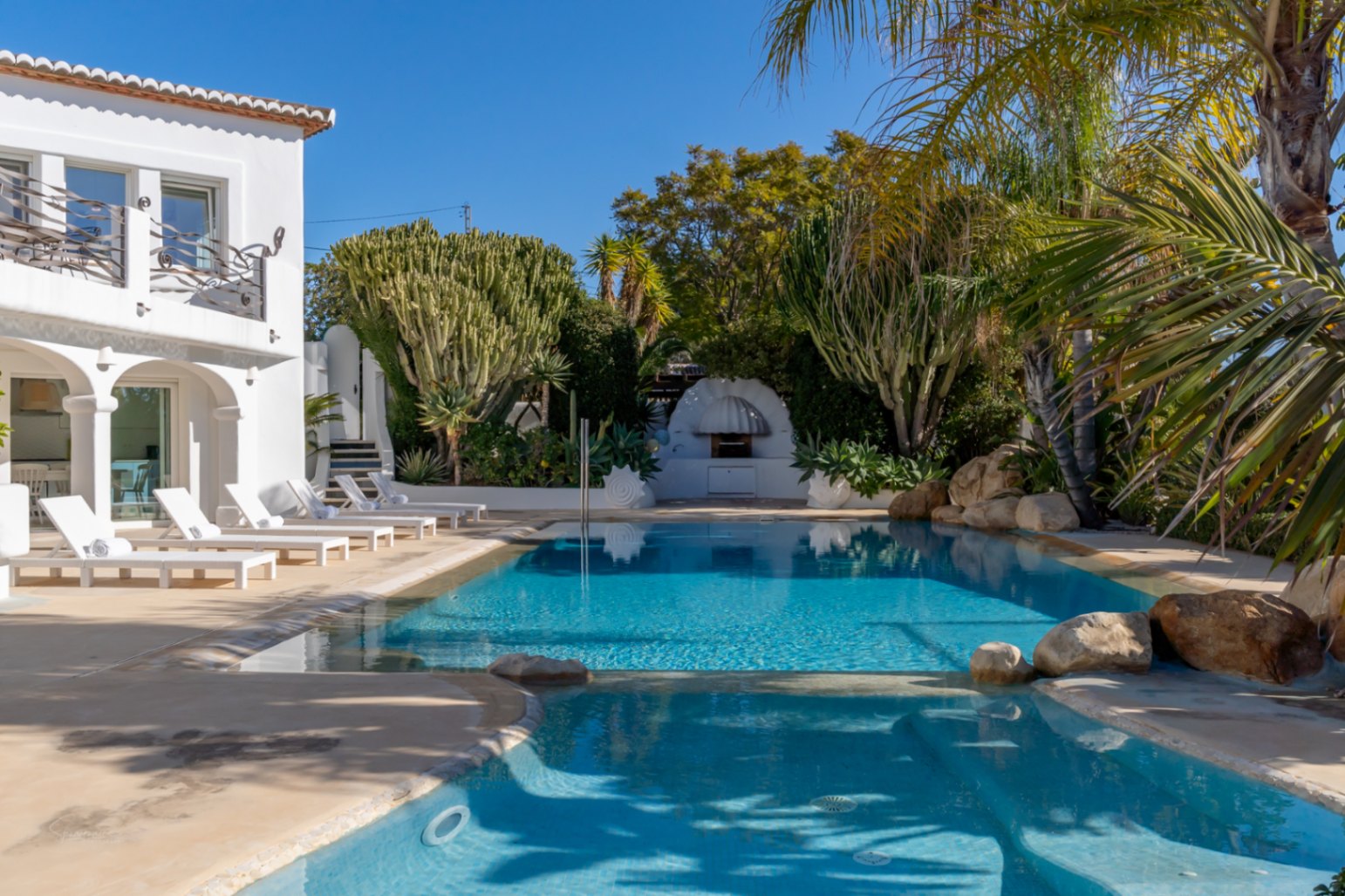 Designer-Pool mit Hydrotherapie-Bereich, weißen Sonnenliegen und üppiger mediterraner Bepflanzung vor der weißen Villa im Ibiza-Stil mit Rundbögen.