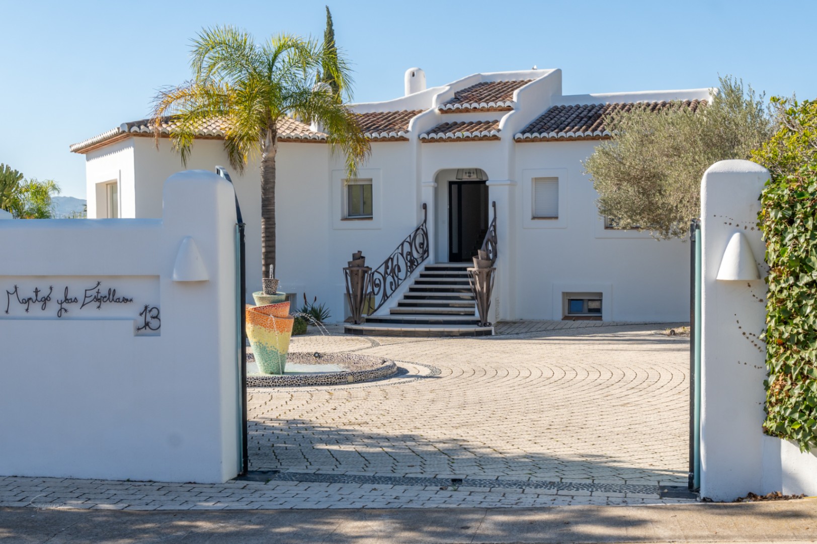 Eingangsbereich im Ibiza-Stil mit weißer Fassade, einem Mosaikbrunnen und gepflastertem Hof. Kunstvolle Schmiedeeisengeländer führen zur erhöhten Haustür.