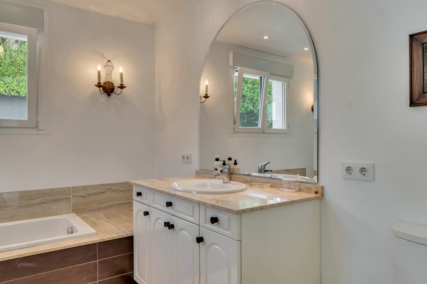 Elegantes Badezimmer mit Marmor-Arbeitsplatte, integriertem Waschbecken und großem Rundbogenspiegel. Mit Einbaubadewanne, Steinoberflächen und klassischen Wandleuchten.