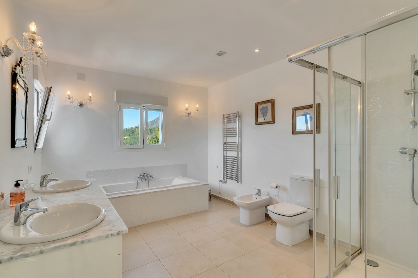 En-Suite-Badezimmer mit Doppelwaschtisch aus Marmor, Einbaubadewanne mit Blick auf den Montgó, Glasdusche und klassischen Wandleuchtern auf hellem Fliesenboden.