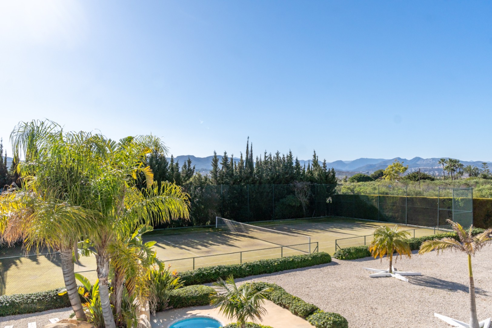 Erhöhter Blick auf den privaten Tennisplatz, umgeben von Hecken und Palmen, mit Aussicht auf das Tal und die Berge unter blauem Himmel.