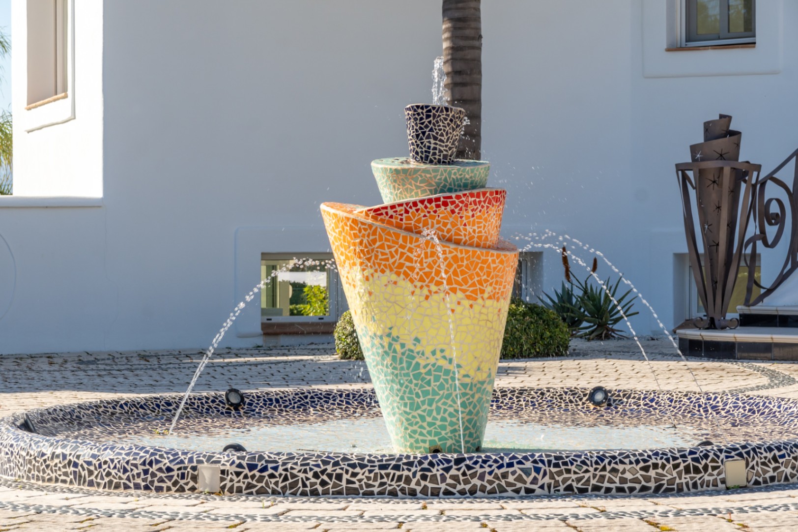 Farbenfroher Mosaikbrunnen im Spiraldesign als zentrales Element im gepflasterten Eingangshof der Villa mit mediterranem Flair.