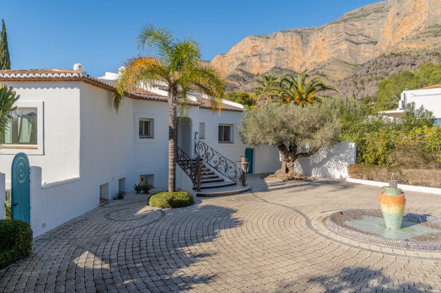 Gepflasterter Eingangshof mit Mosaikbrunnen und Blick auf den Montgó. Weiße Fassade im Ibiza-Stil mit schmiedeeisernen Geländern, Palmen und einem Olivenbaum.