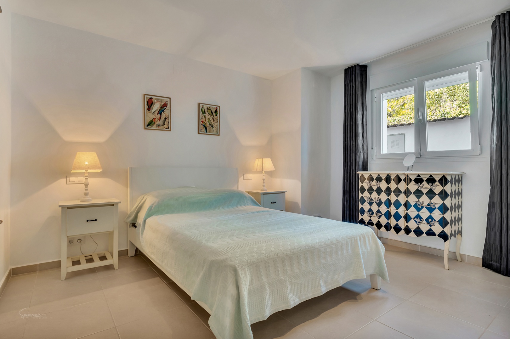 Hell eingerichtetes Schlafzimmer mit Fliesenboden, Doppelbett, weißen Nachttischen und einer markanten Kommode mit Rautenmuster am Fenster.