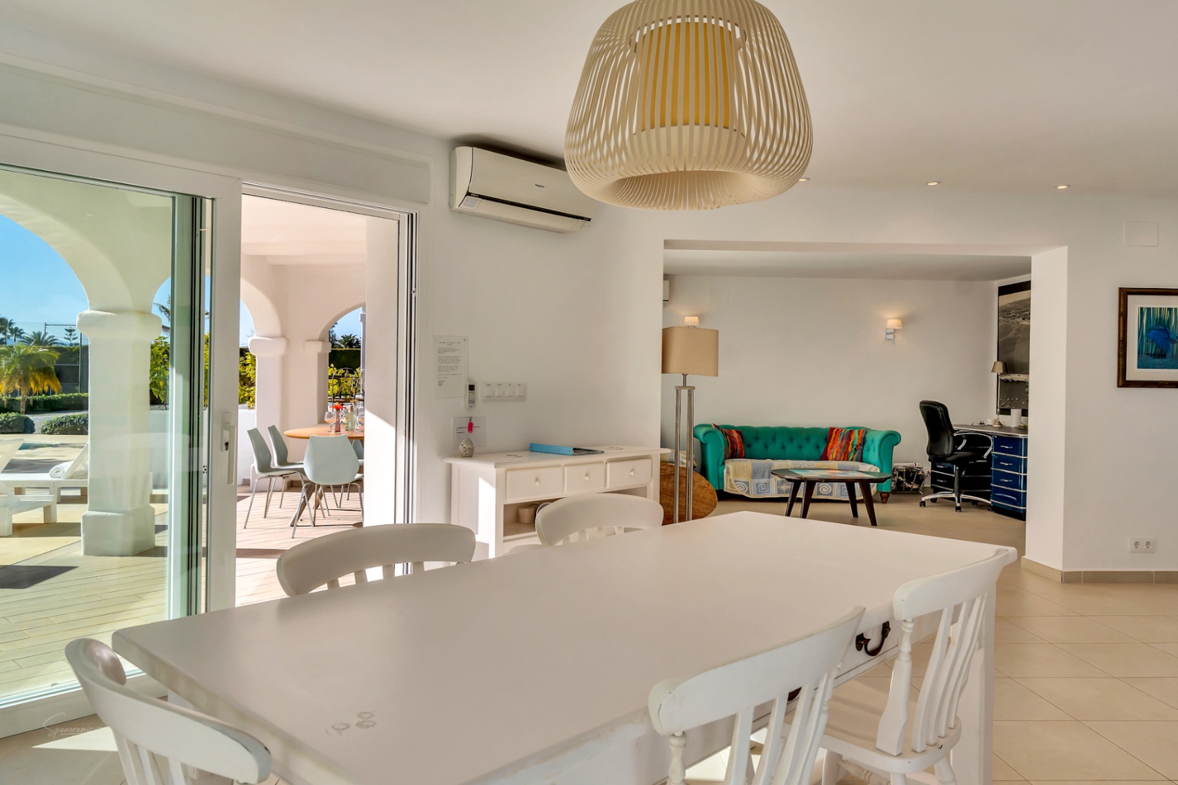 Heller Essbereich mit weißem Holztisch und Korbpendelleuchte, angrenzend an ein Wohnzimmer mit türkisem Sofa, Arbeitsbereich und Zugang zur überdachten Terrasse.