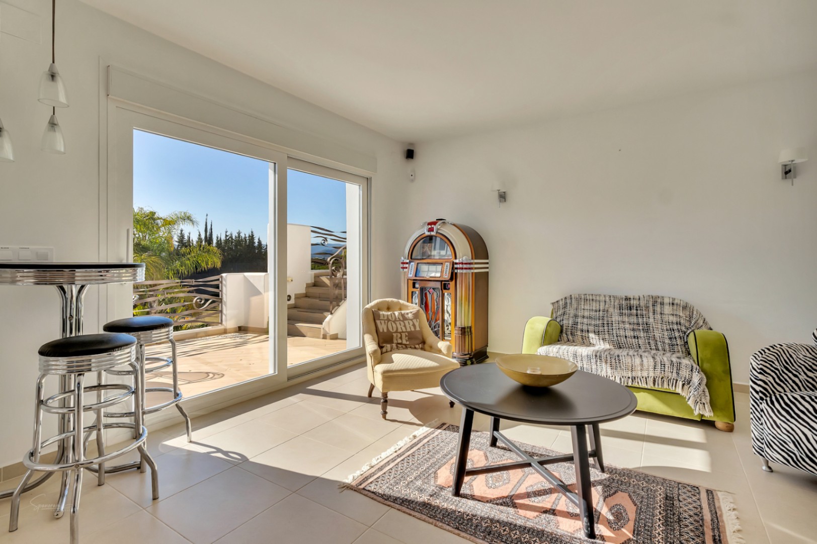 Heller Wohnbereich mit klassischer Jukebox, vielseitigen Sitzmöbeln und großen Glasschiebetüren, die auf eine sonnige Terrasse mit Gartenblick führen.