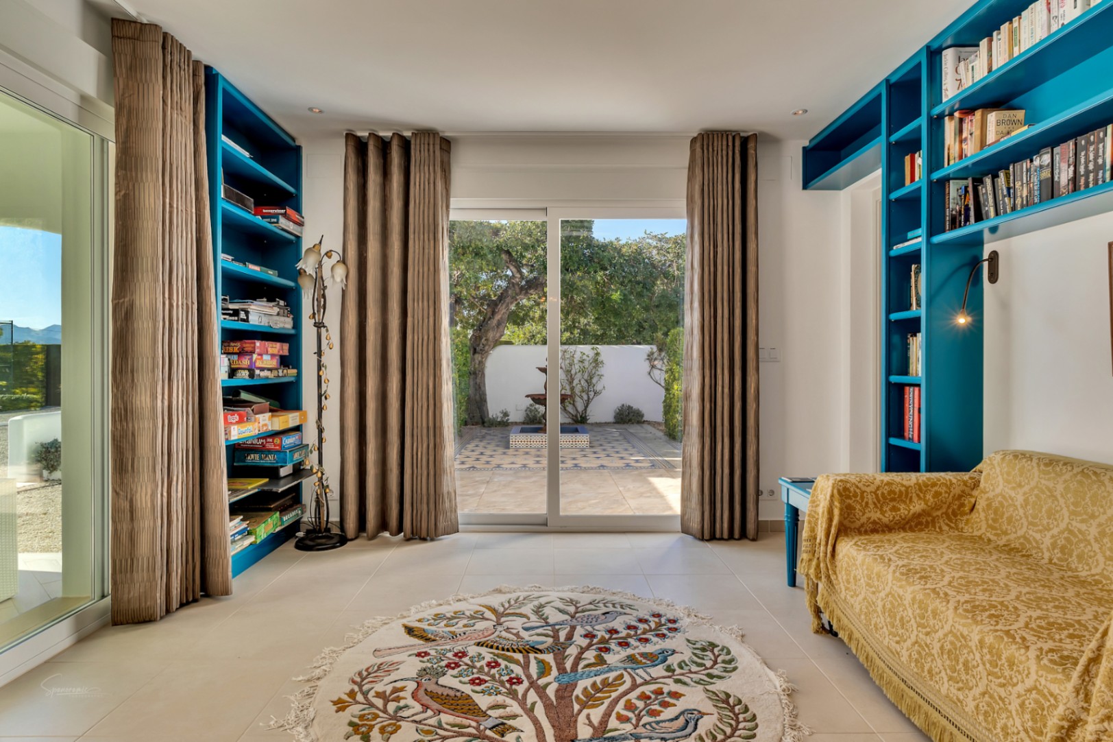 Helles Arbeitszimmer mit blauen Einbauregalen, gelbem Muster-Sofa und Glasschiebetüren zu einem privaten Innenhof mit Springbrunnen und Ziermosaiken.