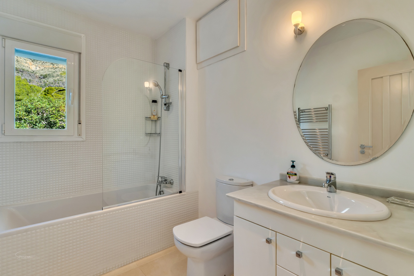 Helles Badezimmer mit weißen Mosaikfliesen, Badewanne mit Glastrennwand und Marmorwaschtisch. Das Fenster bietet einen direkten Blick auf den Berg Montgó.